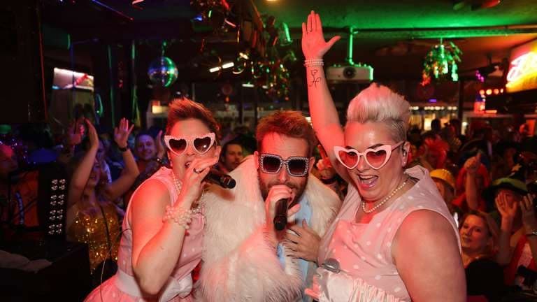 Drei singende Performer mit Herzbrillen vor feiernden Gästen